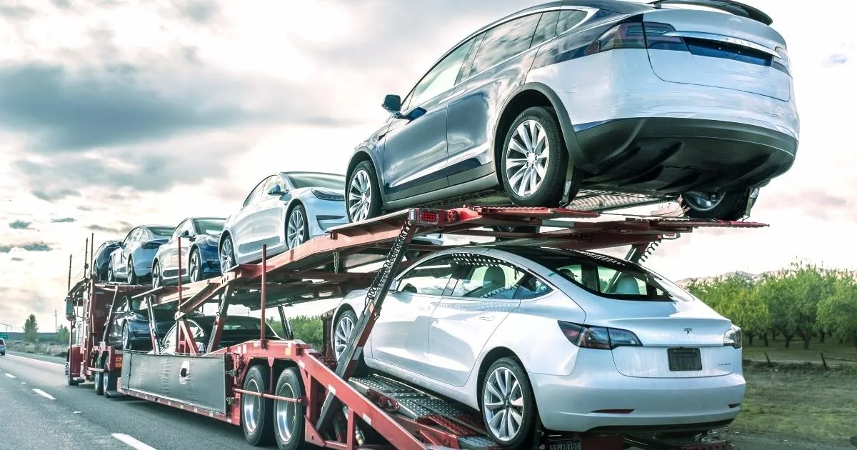 A fleet of silver and white Tesla electric vehicles on a red double-decker car transporter truck.