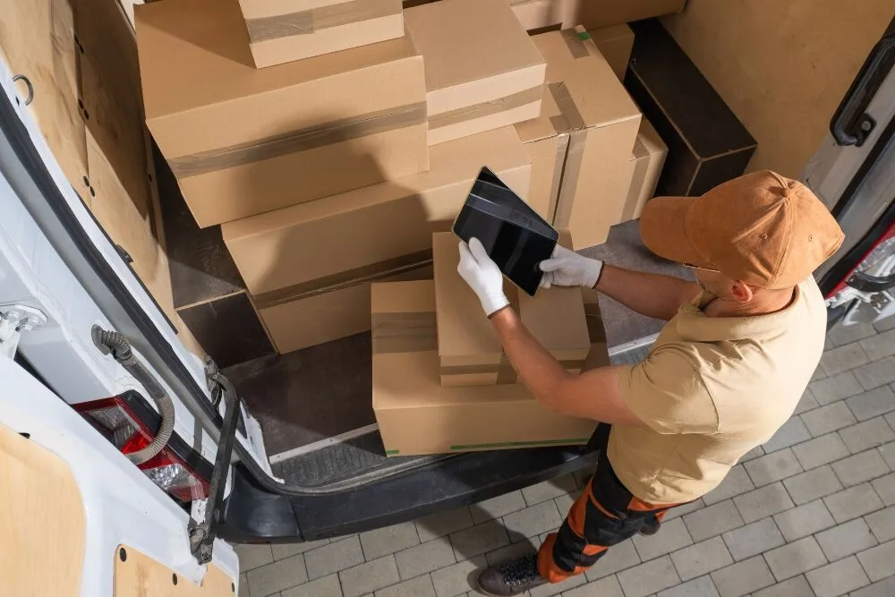 A high-angle view of a delivery worker in a tan uniform and cap standing at the back of a van.