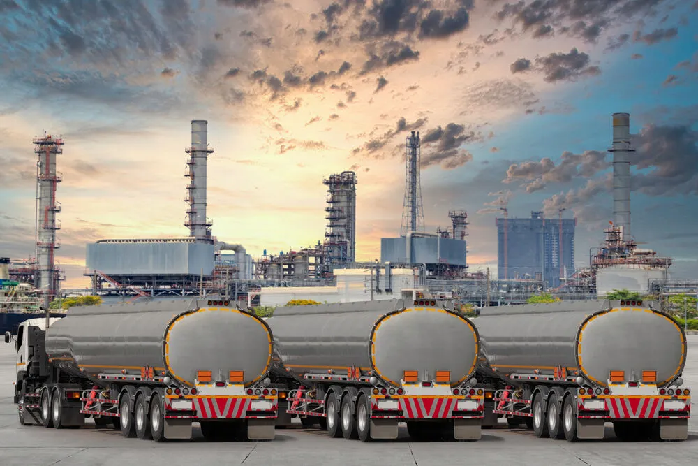 Three silver fuel tanker trucks parked in a row at an industrial refinery