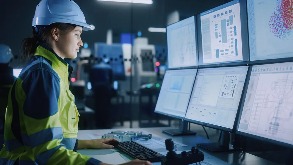 A female engineer in a white hard hat and high-visibility safety jacket sits at a workstation, monitoring complex technical data and schematics across multiple computer screens.