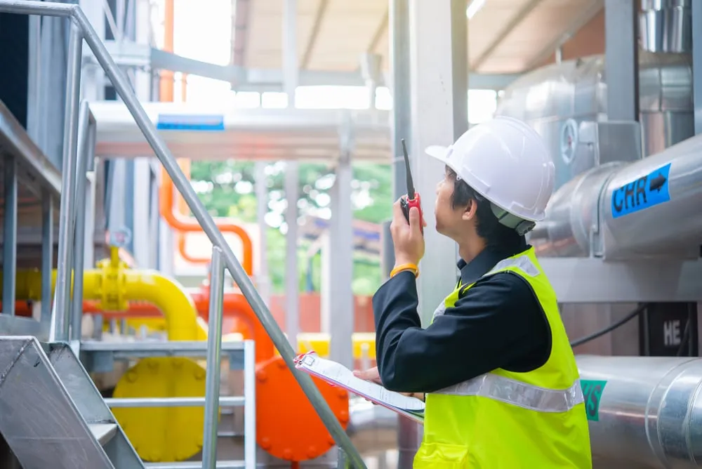 An engineer wearing a white hard hat and a high-visibility yellow vest speaks into a walkie-talkie while holding a clipboard