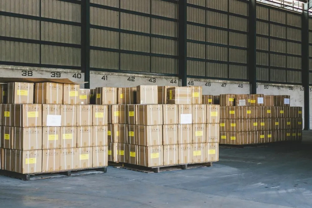Stacks of identical cardboard boxes on wooden pallets are lined up inside a large warehouse