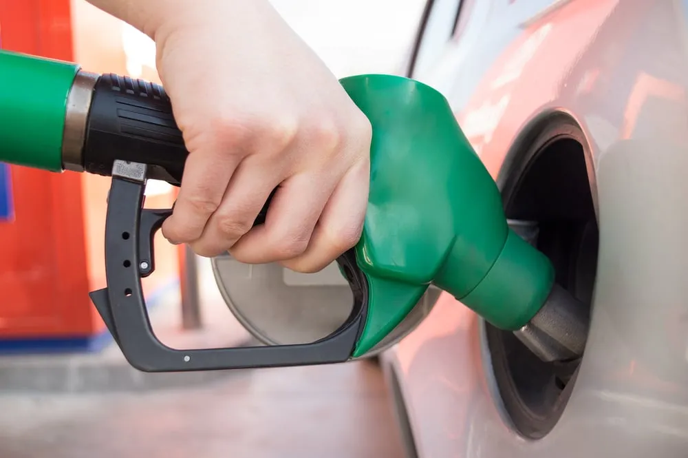 A close-up of a hand holding a green fuel nozzle while refueling a silver car at a gas station.