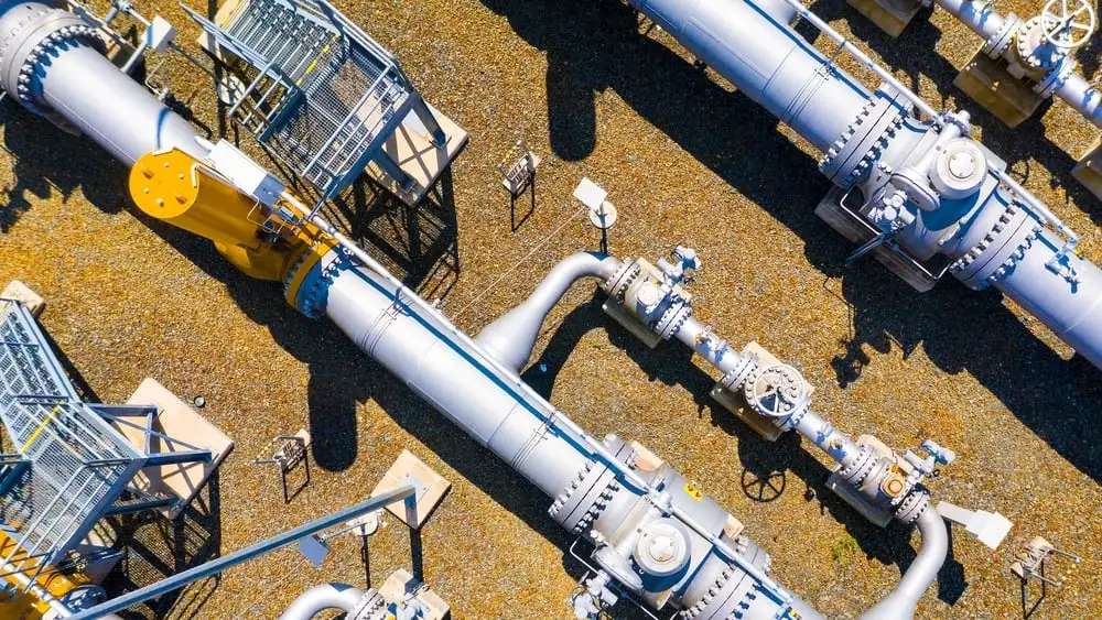An aerial, top-down view of a complex network of large silver industrial pipes and yellow valve assemblies at a facility