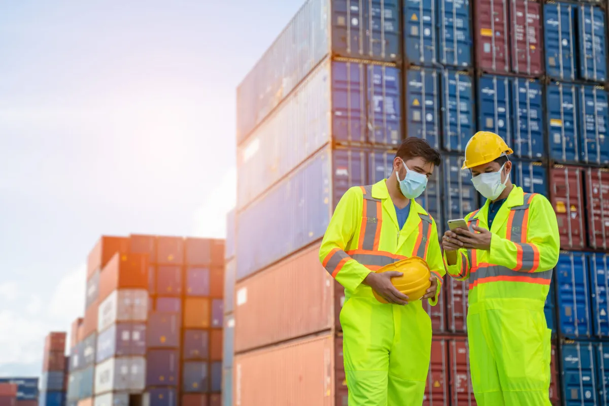 Two logistics workers wearing high-visibility safety coveralls and face masks stand in a shipping yard