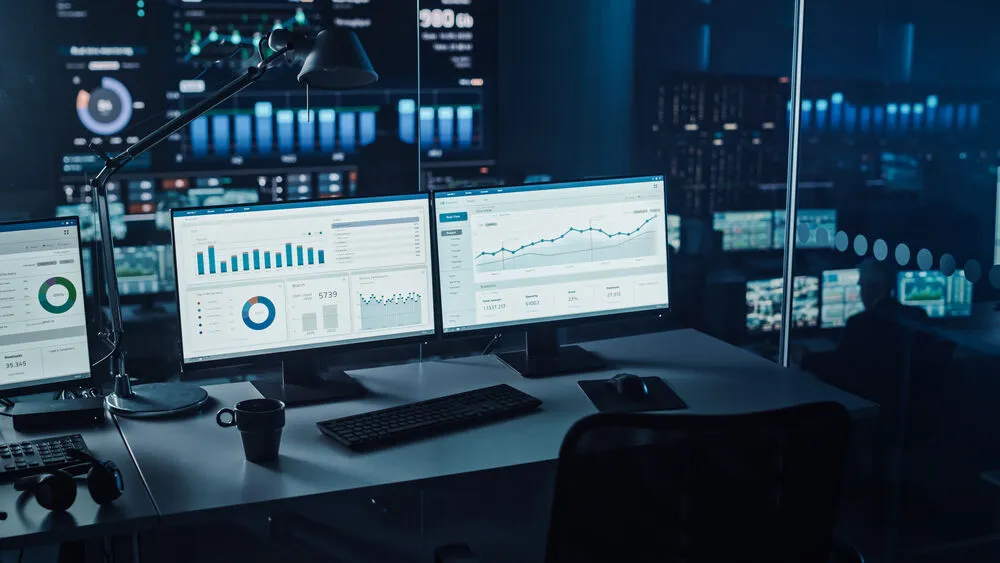 A dark, high-tech control room workstation with dual monitors displaying various business analytics, line graphs, and pie charts.