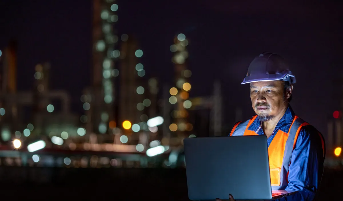 An industrial worker in a white hard hat and orange safety vest looks focused while using a laptop at night