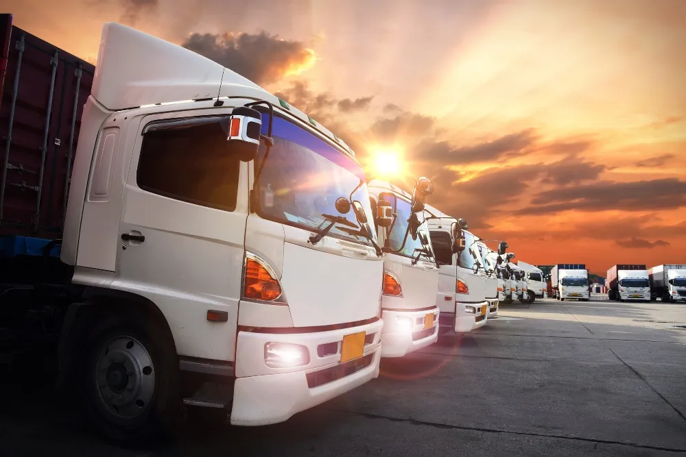 A fleet of white semi-trucks parked in a row at a distribution centre during a vibrant sunset.