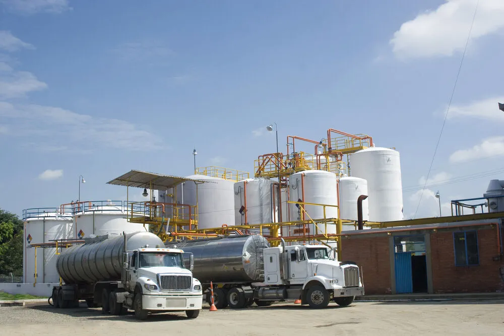 Two white semi-trucks pulling large silver tanker trailers parked at an industrial chemical terminal