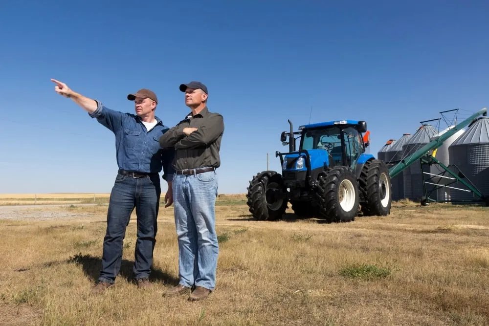 Two farmers stand in a dry field under a clear blue sky.