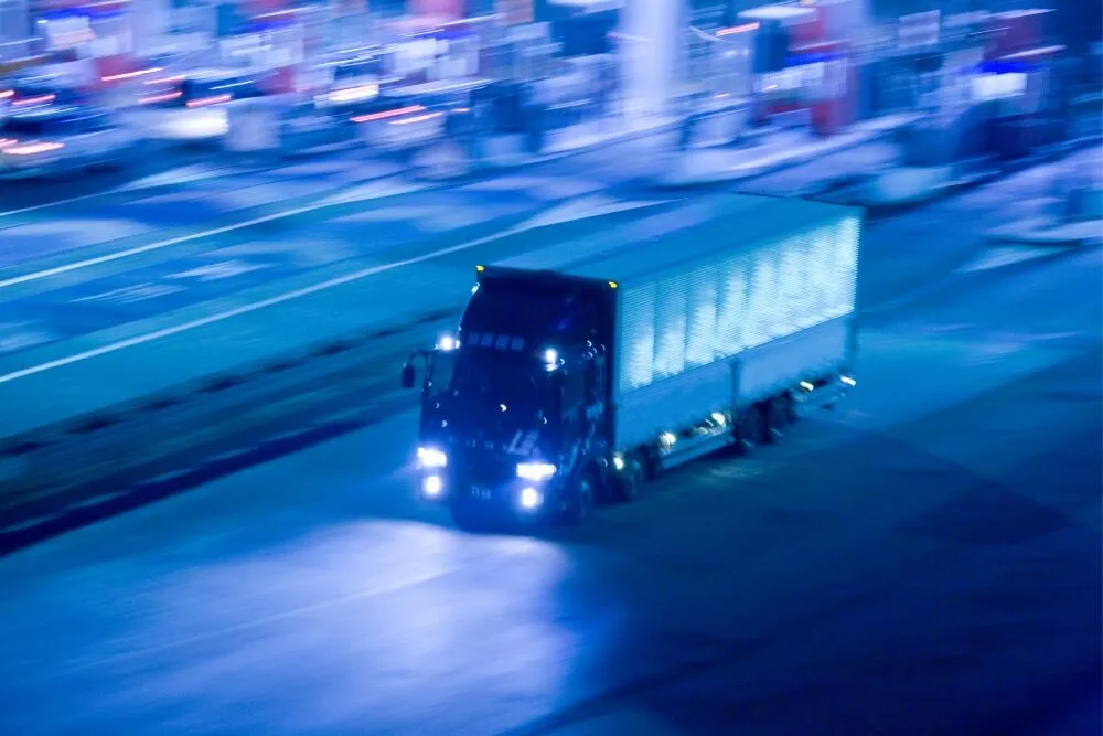 A blue-tinted, long-exposure shot of a delivery truck driving at night with its headlights on.