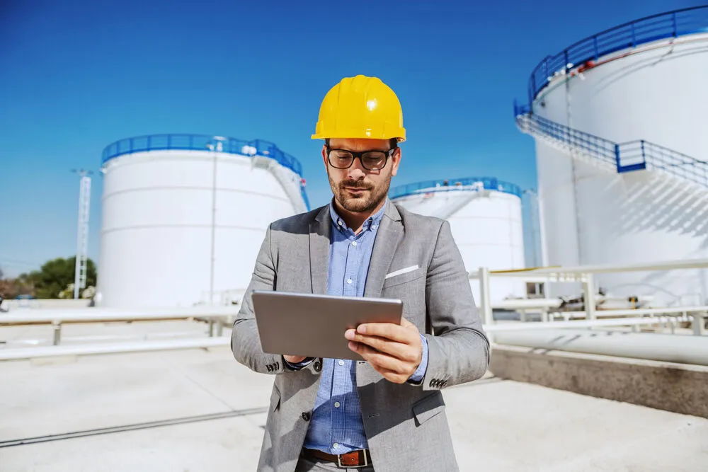 A male engineer in a grey suit and yellow hard hat stands in front of several large, white industrial storage tanks