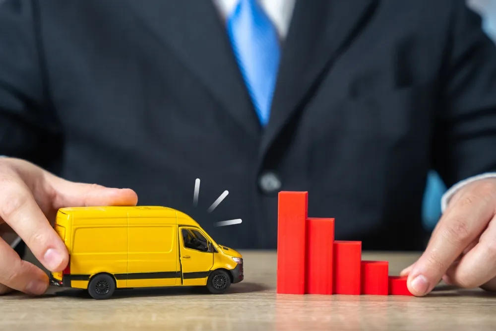 A person in a business suit placing a yellow toy delivery van next to a red toy bar chart that displays a decreasing downward trend on a wooden table.
