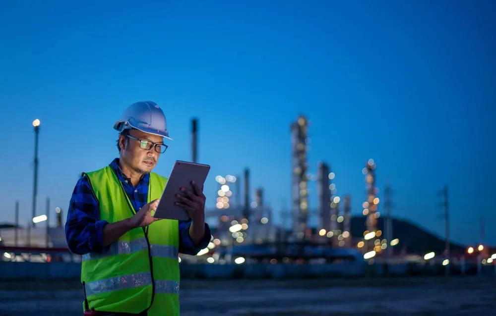 An engineer in a white hard hat and neon safety vest uses a tablet at twilight, with the blurred lights of an industrial refinery glowing in the background.