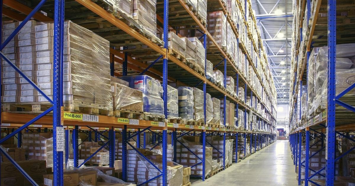 A view down a warehouse aisle lined with tall blue and orange industrial shelving units heavily loaded with shrink-wrapped pallets and inventory boxes.