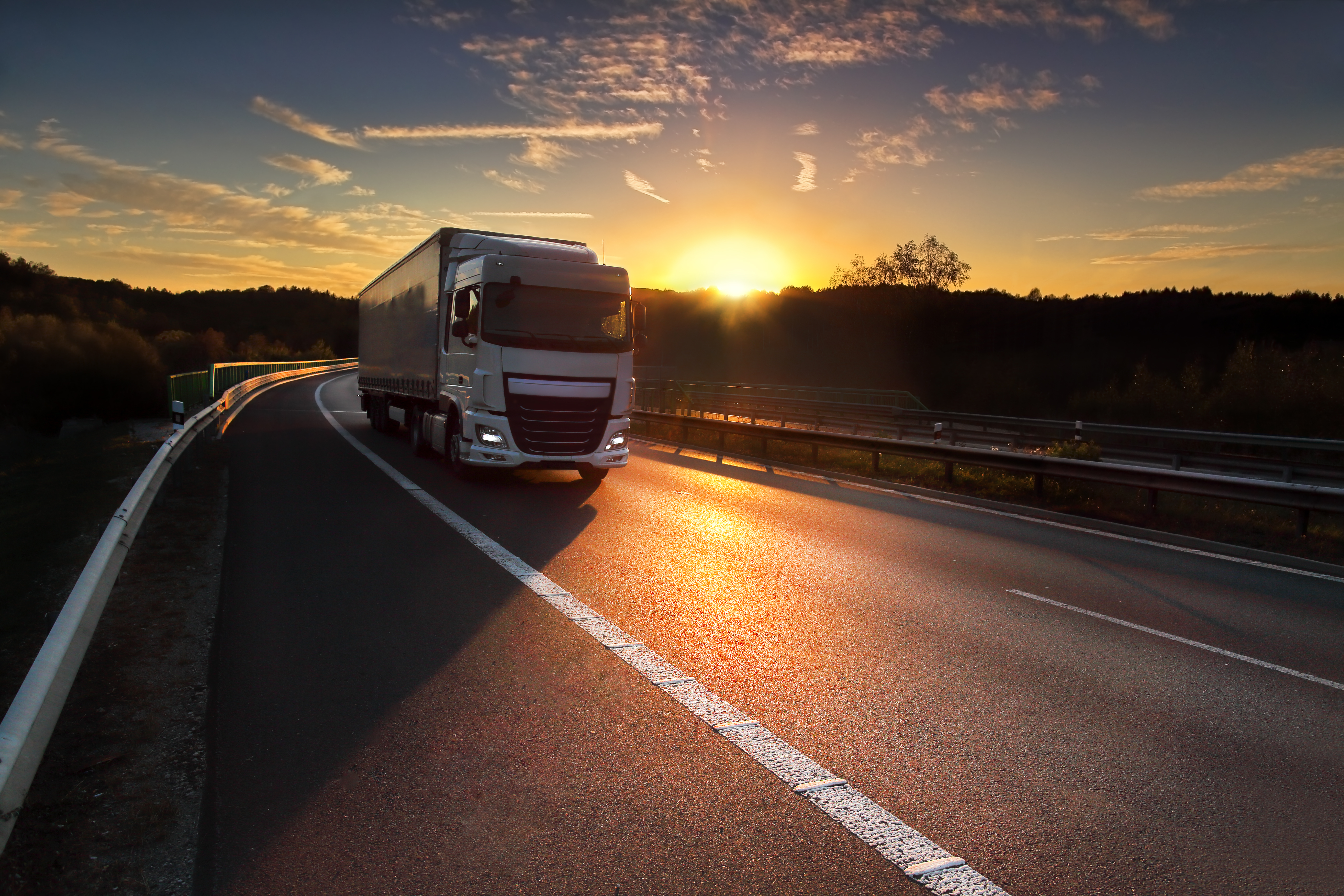 A white semi-truck travels along a winding highway during a vibrant sunset, with golden light reflecting off the road surface.