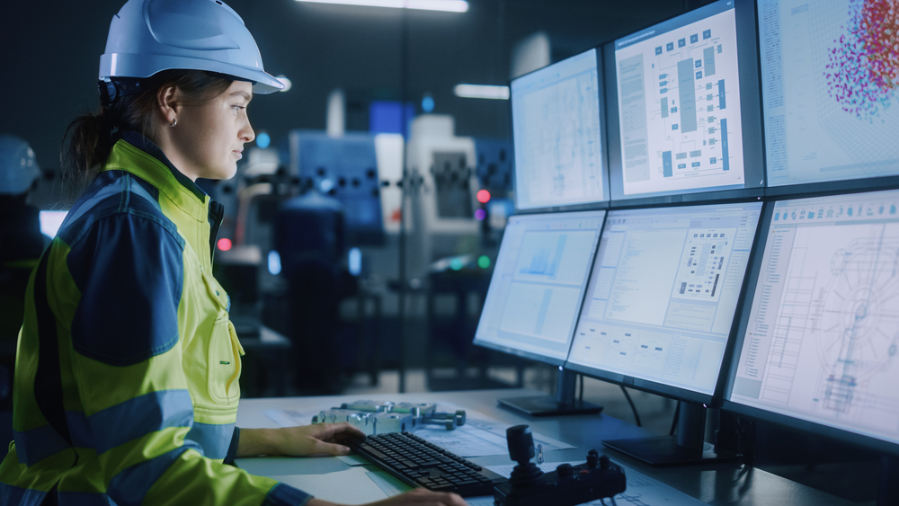 A female engineer in a white hard hat and high-visibility safety jacket sits at a workstation, monitoring complex technical data and schematics across multiple computer screens.