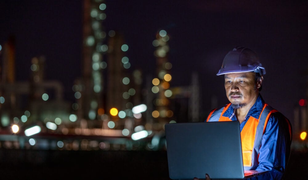 An industrial worker in a white hard hat and orange safety vest looks focused while using a laptop at night