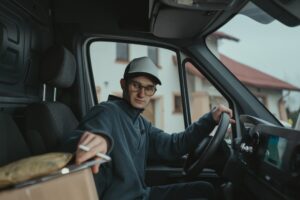 A delivery driver wearing a white cap and glasses sits in the driver's seat of a van