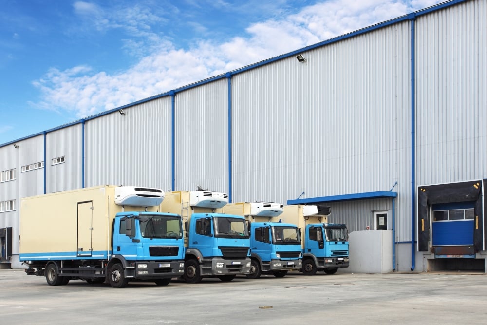 A fleet of four blue-and-tan refrigerated delivery trucks parked in a row outside a large, corrugated metal warehouse.